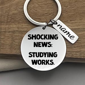 May include: A silver keyring with a round pendant inscribed with "SHOCKING NEWS: STUDYING WORKS." A smaller rectangular tag is attached, with the word "name" engraved. The keyring is displayed on a wooden surface.