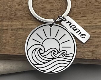 May include: A silver-toned metal keychain featuring a circular charm engraved with a black line art design of a sun rising over ocean waves. Attached to the split ring is a small rectangular metal tag with the word name engraved in script.