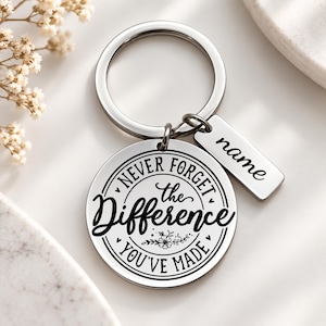 May include: A silver-toned metal keychain featuring a circular pendant engraved with the text "NEVER FORGET the Difference YOU'VE MADE" and a small floral graphic. Attached is a rectangular metal tag with the word "name" engraved on it.