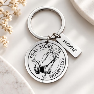 May include: A silver-toned metal keyring featuring a circular pendant with an engraved illustration of praying hands holding a rosary. The text PRAY MORE WORRY LESS is inscribed around the image. A small rectangular tag with the word name is attached.