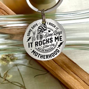 May include: Silver keychain with a black and white design featuring a skeleton with a heart and the text "Some Days I Rock It Some Days It Rocks Me Either Way We're Rockin' Motherhood".