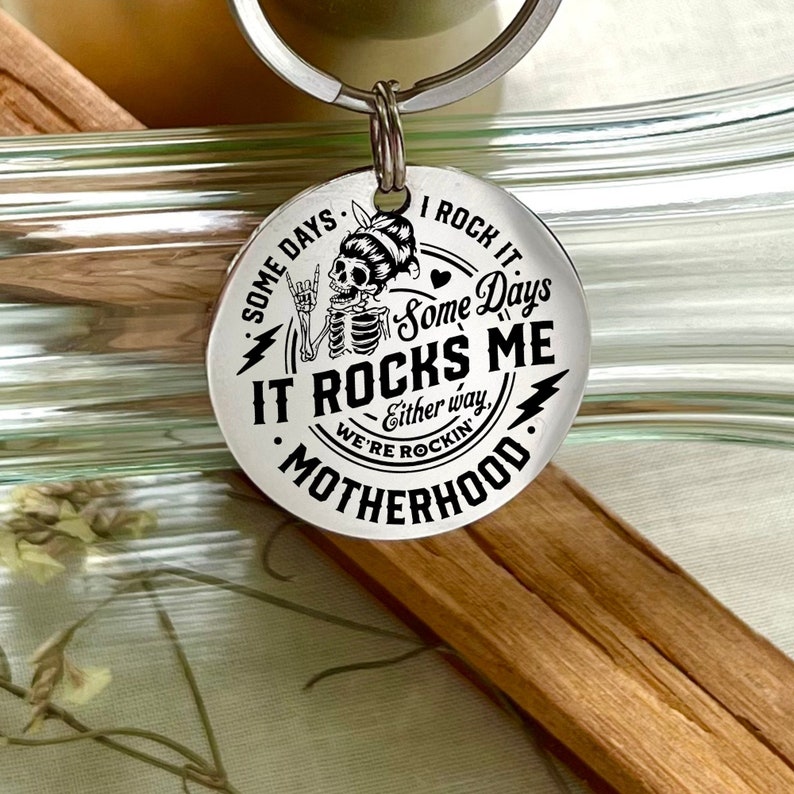 May include: Silver keychain with a black and white design featuring a skeleton with a heart and the text "Some Days I Rock It Some Days It Rocks Me Either Way We're Rockin' Motherhood".