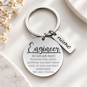 May include: A silver-toned metal keychain featuring a circular pendant engraved with the definition of an engineer, including the text 'Engineer (n) [en-juh-neer] Someone who solves problems you don't know exist, in ways you don't understand. See also: Genius', and a small rectangular tag engraved with the word 'name'.