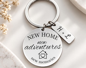 May include: A silver-toned metal keychain featuring a circular disc and a small rectangular tag. The circular disc is engraved with the text NEW HOME new adventures new memories and a house icon with a heart. The rectangular tag displays H + L.