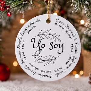 Puede incluir: Un adorno redondo de cerámica blanca con un cordón dorado para colgar. El centro muestra el texto Yo soy rodeado por una corona de hojas negras. El borde incluye frases en español como Siempre Amada, Capaz, Nunca Sola y versículos bíblicos.