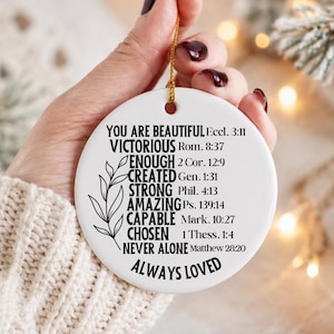 Può includere: Un ornamento rotondo in ceramica bianca con testo nero: YOU ARE BEAUTIFUL Eccl. 3:11, VICTORIOUS Rom. 8:37, ENOUGH 2 Cor. 12:9, CREATED Gen. 1:31, STRONG Phil. 4:13, AMAZING Ps. 139:14, CAPABLE Mark. 10:27, CHOSEN 1 Thess. 1:4, NEVER ALONE Matthew 28:20, ALWAYS LOVED. Un disegno a linee nere di un ramo frondoso si trova sul lato sinistro e un cordino dorato è attaccato alla parte superiore.