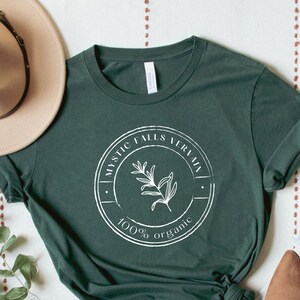May include: A dark green t-shirt with a white circle graphic that says "Mystic Falls Verbena 100% organic" with a small plant illustration inside the circle.
