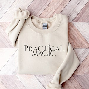 May include: A beige sweatshirt with the text "PRACTICAL MAGIC" printed in black on the front. A small star is above the word "MAGIC".