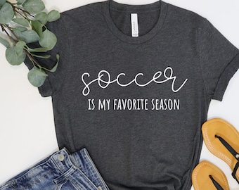 Könnte beinhalten: Ein dunkelgraues T-Shirt mit dem Text "soccer is my favorite season" in weißen Buchstaben.