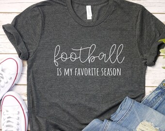 May include: Dark gray t-shirt with the text "football is my favorite season" in white script. The shirt is paired with blue jeans and white canvas sneakers. The shirt is folded, and the jeans are distressed.