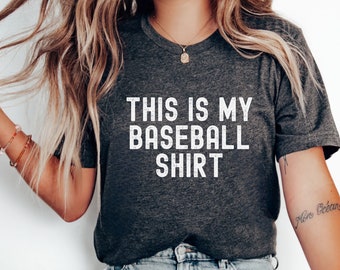 May include: A dark gray t-shirt with the text "This is my baseball shirt" printed in white. The shirt is being worn by a person with long blonde hair and is paired with light blue jeans.