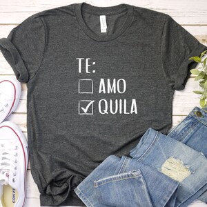 May include: A dark gray t-shirt with a white graphic that says "TE:  AMO  QUILA" with a checkmark next to "QUILA".