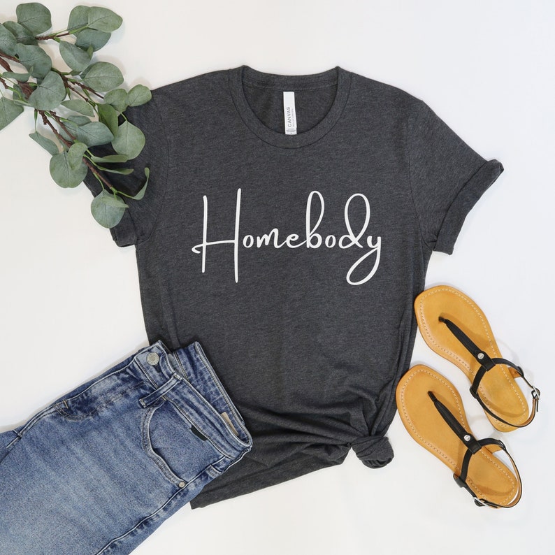 Puede incluir: Una camiseta gris oscuro con la palabra "Homebody" escrita en cursiva blanca. La camiseta est&aacute; colocada sobre una superficie blanca con un par de sandalias marrones y unos vaqueros azules.