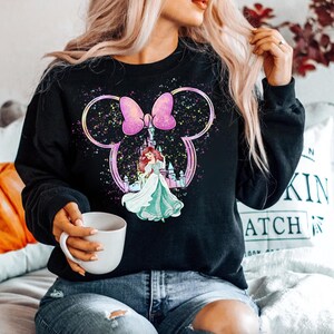 Puede incluir: Sudadera negra con un lazo rosa y blanco y un gr&aacute;fico de Ariel de La Sirenita frente al castillo de Cenicienta. El gr&aacute;fico est&aacute; rodeado de un fondo brillante y brillante.