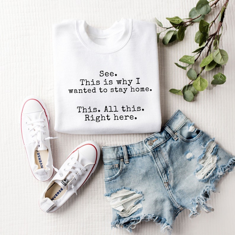 May include: White sweatshirt with the text "See. This is why I wanted to stay home. This. All this. Right here." printed on it. The sweatshirt is laid out on a white surface with a pair of white sneakers and denim shorts.