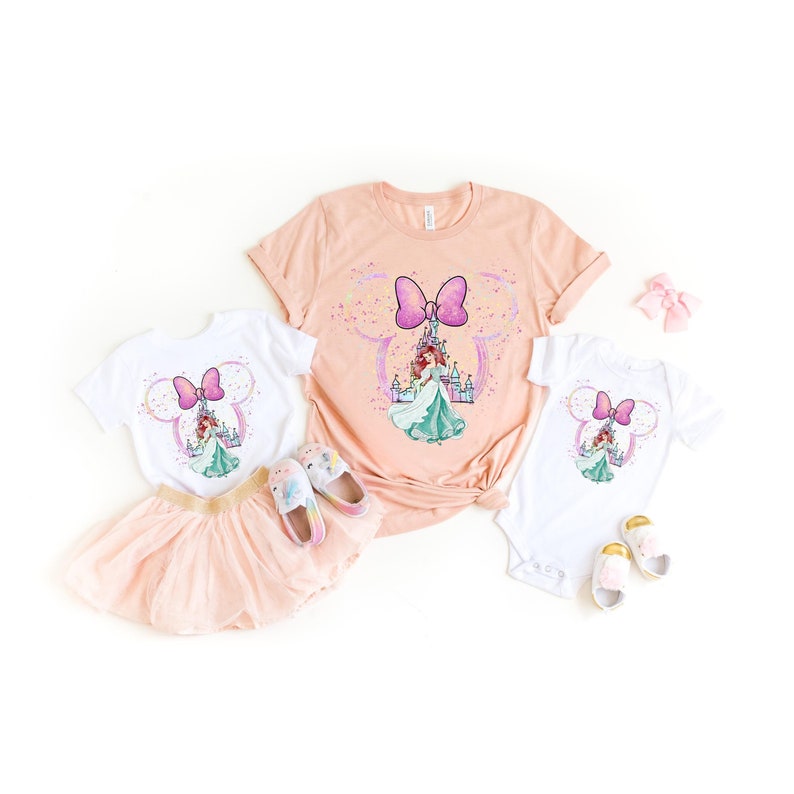 Puede incluir: Un conjunto de dos camisetas blancas con un lazo rosa y una princesa frente a un castillo. La camiseta m&aacute;s grande es de color melocot&oacute;n y la m&aacute;s peque&ntilde;a es un body. Las camisetas est&aacute;n dispuestas sobre un fondo blanco.