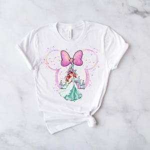 Puede incluir: Camiseta blanca con un lazo rosa y una ilustraci&oacute;n de acuarela de una princesa frente a un castillo. La ilustraci&oacute;n est&aacute; rodeada por una forma de oreja de Mickey Mouse con un fondo brillante.