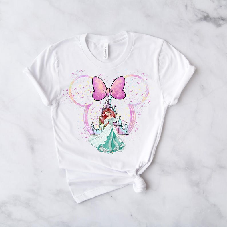 Puede incluir: Camiseta blanca con un lazo rosa y una ilustraci&oacute;n de acuarela de una princesa frente a un castillo. La ilustraci&oacute;n est&aacute; rodeada por una forma de oreja de Mickey Mouse con un fondo brillante.