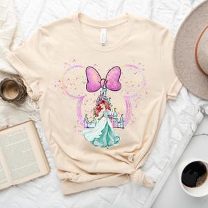 Puede incluir: Una camiseta beige claro con un lazo rosa y una ilustraci&oacute;n de acuarela de Ariel de La Sirenita frente al castillo de Cenicienta. La ilustraci&oacute;n est&aacute; rodeada por una silueta de oreja de Mickey Mouse de color rosa y azul claro.