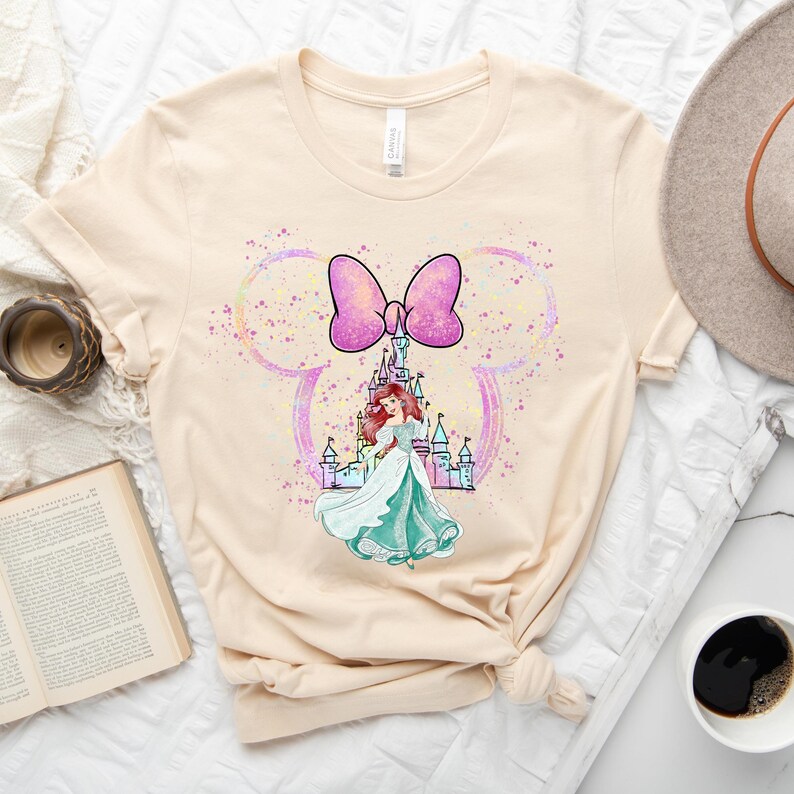 Puede incluir: Una camiseta beige claro con un lazo rosa y una ilustraci&oacute;n de acuarela de Ariel de La Sirenita frente al castillo de Cenicienta. La ilustraci&oacute;n est&aacute; rodeada por una silueta de oreja de Mickey Mouse de color rosa y azul claro.