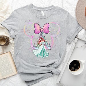 Puede incluir: Una camiseta gris con un lazo rosa y una princesa de pie frente a un castillo. La princesa lleva un vestido verde y tiene el pelo largo y rojo. El castillo est&aacute; rodeado de purpurina rosa.