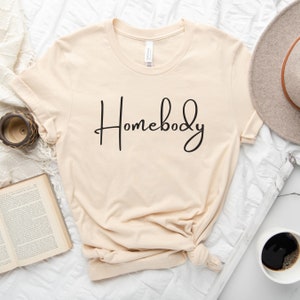 Puede incluir: Una camiseta beige con el texto "Homebody" escrito en cursiva negra.