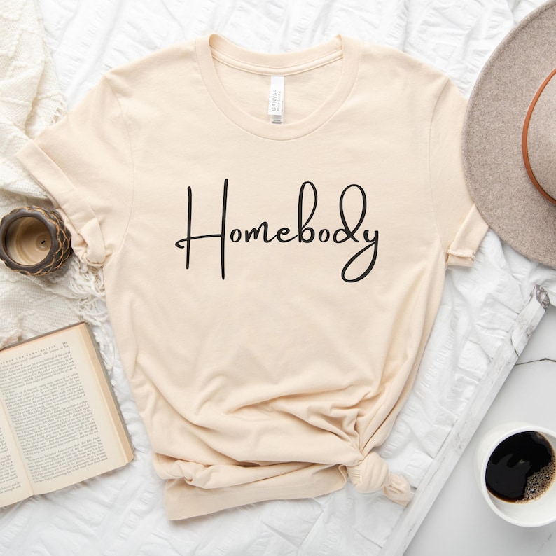 Puede incluir: Una camiseta beige con el texto "Homebody" escrito en cursiva negra.