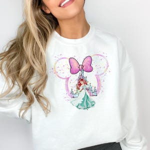 Puede incluir: Sudadera blanca con un lazo rosa y morado y una imagen de dibujos animados de Ariel de La Sirenita frente a un castillo rosa. La imagen est&aacute; rodeada por un contorno blanco de las orejas de Mickey Mouse.