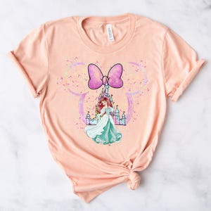 Puede incluir: Camiseta de color melocot&oacute;n con un lazo rosa, un castillo y una princesa con un vestido verde. El dise&ntilde;o est&aacute; rodeado de purpurina.