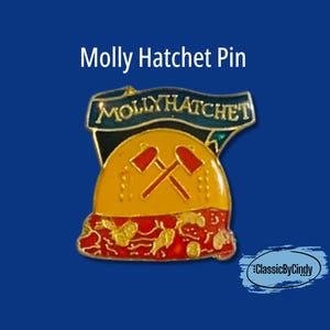 May include: A Molly Hatchet pin with a gold, red, and blue design. The pin features the band's name in a banner above a yellow shape with crossed pickaxes. The bottom has a red and gold design.