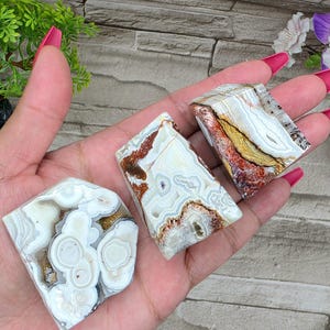 May include: Three polished agate stones with unique patterns of white, brown, and gold. The stones are held in a hand, showcasing their natural textures and colors. The agate pieces vary in shape and size, highlighting their individual characteristics.