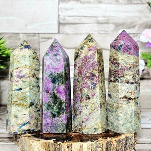 May include: Four polished gemstone towers with a variety of colors, including pink, green, and beige. The towers are displayed on a wooden base, showcasing their unique patterns and textures. Each tower has a pointed top.