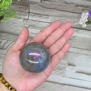 May include: A polished, iridescent sphere with a mix of blue, purple, and green hues. The gemstone ball is held in an open hand, showcasing its smooth, rounded form. The sphere's surface reflects light, creating a shimmering effect.
