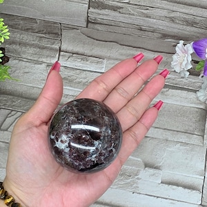 May include: A polished, dark red sphere with a marbled appearance. The gemstone ball is held in a hand, showcasing its smooth, reflective surface. The sphere's color ranges from deep burgundy to lighter shades, creating a natural, patterned effect. The background is a gray brick wall.