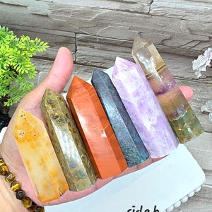 May include: A collection of six polished crystal points held in a hand, featuring a yellow stone, a speckled olive-green stone, a smooth orange stone, a dark blue-grey stone, a light purple stone, and a multi-colored translucent stone labeled side b.