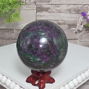 May include: A polished, spherical ruby in zoisite gemstone featuring deep purple and dark green mottled patterns. The stone is displayed on a small, carved, reddish-brown wooden tripod stand, showcasing its smooth, reflective surface and natural mineral inclusions.
