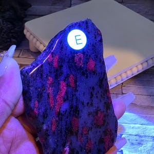 Op de afbeelding: Een gepolijste, onregelmatige steenplaat met een donkere, gevlekte basis en levendige, gloeiende roze-rode insluitingen onder ultraviolet licht. Een kleine, ronde witte sticker met de letter E is op de steen aangebracht.