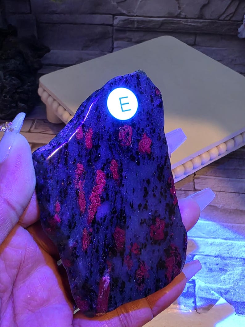 Op de afbeelding: Een gepolijste, onregelmatige steenplaat met een donkere, gevlekte basis en levendige, gloeiende roze-rode insluitingen onder ultraviolet licht. Een kleine, ronde witte sticker met de letter E is op de steen aangebracht.