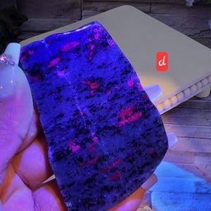 Op de afbeelding: Een platte, onregelmatig gevormde plaat van mineraalgesteente met een donkerblauwe en zwart gespikkelde basis met levendige, gloeiende roze insluitingen. De steen wordt verlicht onder ultraviolet licht, wat de fluorescerende roze patronen benadrukt.
