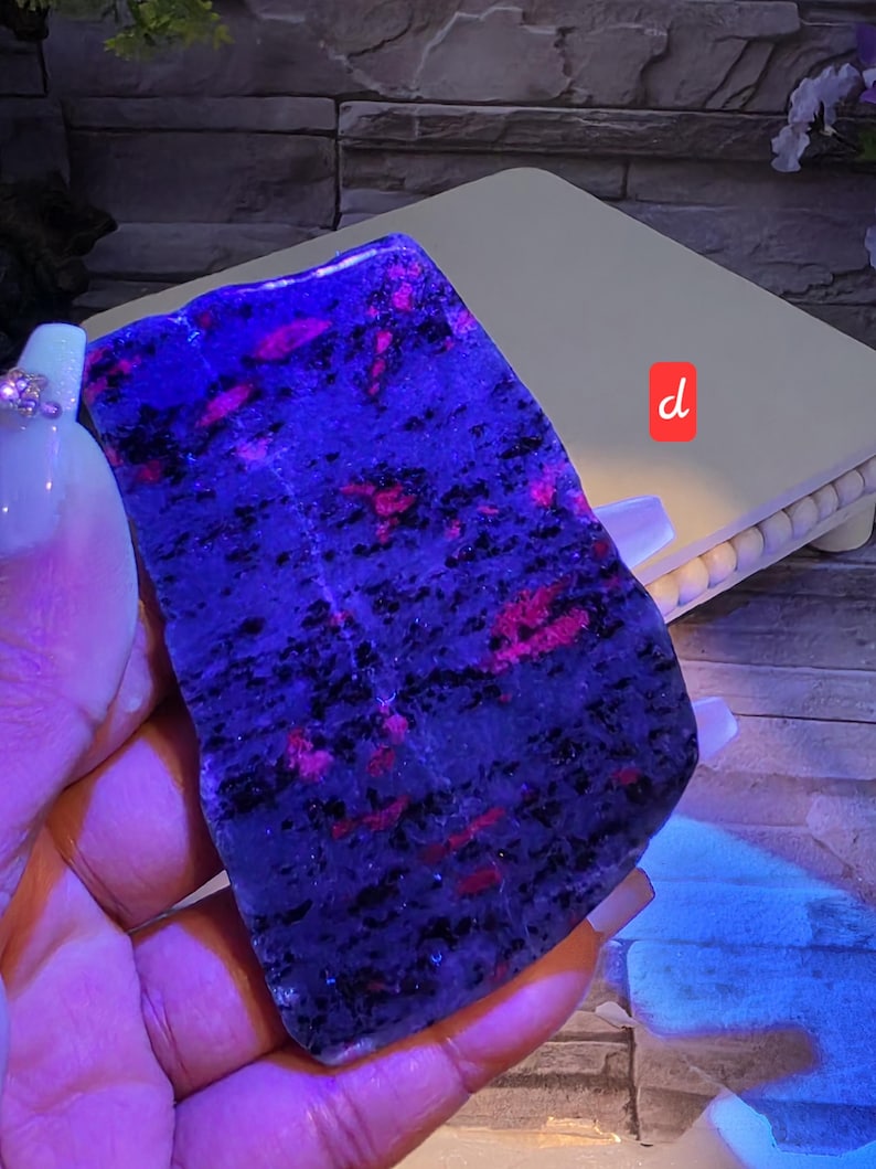 Op de afbeelding: Een platte, onregelmatig gevormde plaat van mineraalgesteente met een donkerblauwe en zwart gespikkelde basis met levendige, gloeiende roze insluitingen. De steen wordt verlicht onder ultraviolet licht, wat de fluorescerende roze patronen benadrukt.