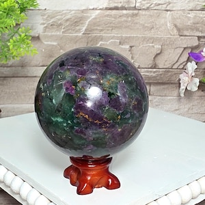 May include: A polished spherical gemstone featuring a mottled pattern of deep purple and dark green mineral inclusions with scattered metallic gold-colored flecks, resting securely on a small, carved, reddish-brown wooden tripod display stand.