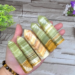 May include: A hand holding several green onyx crystal points. The crystals have a banded pattern with shades of green, white, and gold. The crystals are polished and have a pointed top. The background is a light gray brick wall.