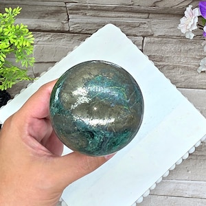 May include: A polished spherical stone featuring a complex marbled pattern of deep forest green, teal, and metallic bronze-gold inclusions. The surface is smooth and reflective, showcasing a natural mineral composition with swirling dark and light tones.