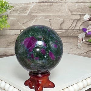 May include: A polished spherical stone featuring a deep green base with vibrant magenta-colored inclusions throughout. The sphere is mounted on a small, glossy, reddish-brown wooden stand with a tripod base, showcasing a smooth, reflective surface.