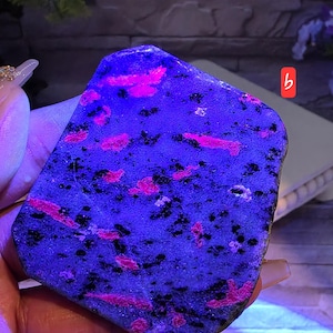 Op de afbeelding: Een platte, onregelmatig gevormde steenplaat met een donkerblauwe basis met verspreide, levendige neonroze insluitingen en kleine zwarte spikkels. Het oppervlak ziet er getextureerd uit en wordt verlicht onder ultraviolet licht, gemarkeerd met een klein rood label.
