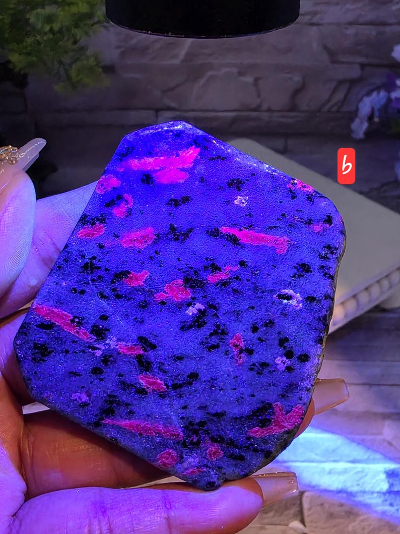 Op de afbeelding: Een platte, onregelmatig gevormde steenplaat met een donkerblauwe basis met verspreide, levendige neonroze insluitingen en kleine zwarte spikkels. Het oppervlak ziet er getextureerd uit en wordt verlicht onder ultraviolet licht, gemarkeerd met een klein rood label.