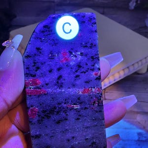 Op de afbeelding: Een rechthoekige plaat van gepolijste steen met een donkergrijze en zwarte gespikkelde basis met levendige rode insluitingen. De steen wordt verlicht onder ultraviolet licht, wat een witte ronde sticker met de letter C op de bovenkant onthult.