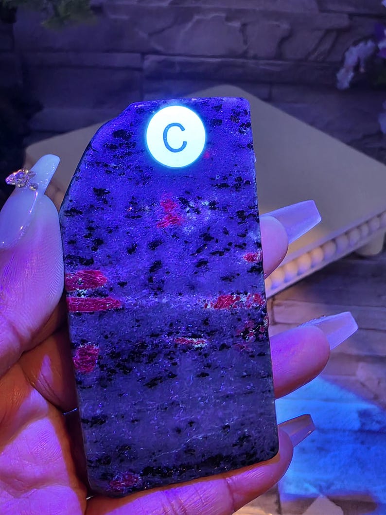 Op de afbeelding: Een rechthoekige plaat van gepolijste steen met een donkergrijze en zwarte gespikkelde basis met levendige rode insluitingen. De steen wordt verlicht onder ultraviolet licht, wat een witte ronde sticker met de letter C op de bovenkant onthult.