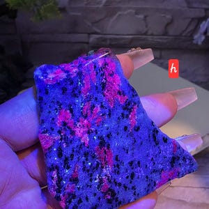 May include: A rough, flat piece of mineral specimen featuring a vibrant, fluorescent pink and deep blue-purple color palette with scattered black inclusions. The surface has a crystalline, textured appearance under ultraviolet light, marked with a small red tag.