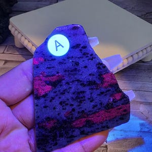 Op de afbeelding: Een platte, onregelmatig gevormde plaat van mineraalsteen met een donkerpaarse en zwarte basis met levendige, gloeiende magentakleurige vlekken. Een ronde witte sticker met de letter A is bevestigd op het bovenste deel van het stenen oppervlak.